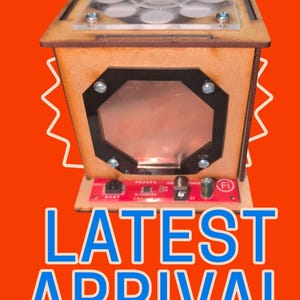 Op de afbeelding: Een houten doos met een doorzichtige bovenkant en een koperkleurig voorpaneel. De doos heeft een printplaatbasis met de tekst "Fi". De woorden "NEW!" worden bovenaan herhaald en "LATEST ARRIVAL" staat in het blauw onderaan, alles tegen een oranje achtergrond.