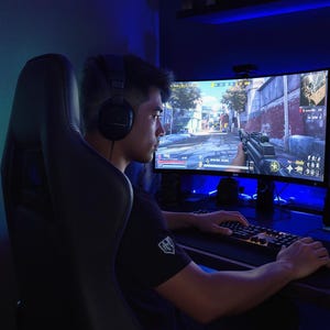 以下が含まれることがあります： ヘッドフォンをつけた人が、ビデオゲームを表示する湾曲したコンピューターモニターに集中しています。その人は黒いゲーミングチェアに座っており、キーボードとマウスが見えます。部屋は青い光で照らされています。