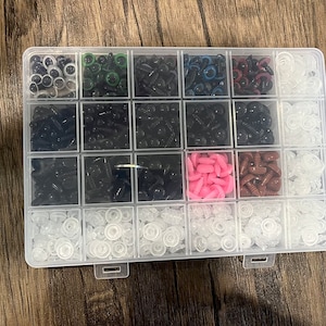 May include: A clear plastic organizer filled with various sizes and colors of craft eyes. The assortment includes black, white, pink, brown, and green eyes, perfect for crafting. The organizer is on a wooden surface.