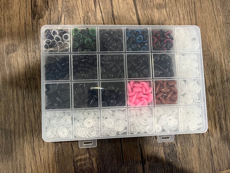 May include: A clear plastic organizer filled with various sizes and colors of craft eyes. The assortment includes black, white, pink, brown, and green eyes, perfect for crafting. The organizer is on a wooden surface.
