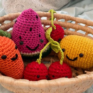 May include: A woven basket filled with colorful, crocheted fruit and vegetables. The collection includes a smiling orange carrot, a watermelon slice, a pair of cherries, and a yellow lemon. Each piece has a smiling face.
