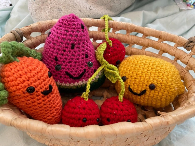 May include: A woven basket filled with colorful, crocheted fruit and vegetables. The collection includes a smiling orange carrot, a watermelon slice, a pair of cherries, and a yellow lemon. Each piece has a smiling face.