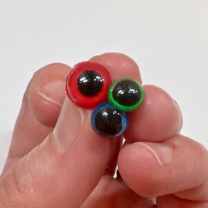 May include: A close-up of three colorful craft googly eyes. The eyes are red, green, and blue with black pupils. The eyes are held in a person's hand against a white background.
