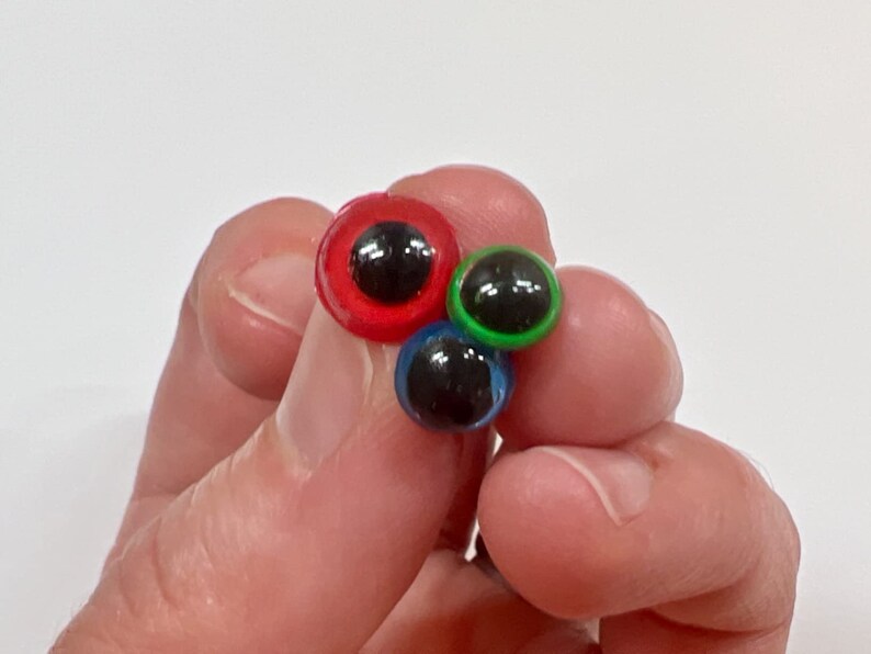 May include: A close-up of three colorful craft googly eyes. The eyes are red, green, and blue with black pupils. The eyes are held in a person's hand against a white background.