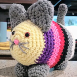 May include: A handmade crochet cat toy with a light yellow head, gray ears, and a striped body in shades of purple, pink, red, black, and white. The cat has pink nose, gray whiskers, and red eyes. The toy is sitting on gray paws.