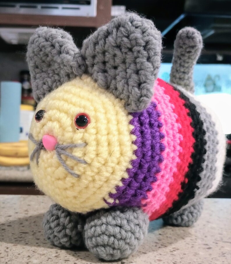 May include: A handmade crochet cat toy with a light yellow head, gray ears, and a striped body in shades of purple, pink, red, black, and white. The cat has pink nose, gray whiskers, and red eyes. The toy is sitting on gray paws.
