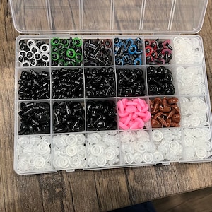 May include: A clear plastic organizer filled with various sizes and colors of craft safety eyes and washers. The eyes are black, white, green, blue, and red. The washers are clear and white. The organizer is on a wooden surface.