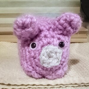 May include: A handmade, lavender-colored crocheted bear with a white and silver snout. The bear has two black plastic eyes and two small, rounded ears. The crochet work has a sparkly, glittery effect.