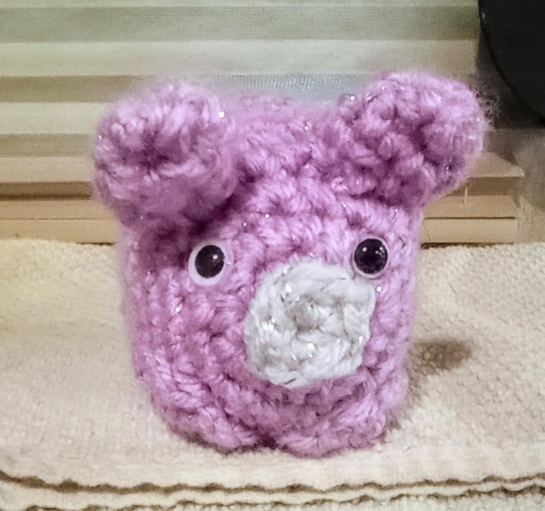 May include: A handmade, lavender-colored crocheted bear with a white and silver snout. The bear has two black plastic eyes and two small, rounded ears. The crochet work has a sparkly, glittery effect.