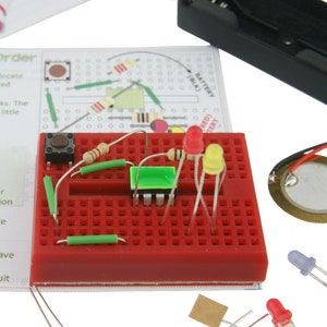 May include: A red breadboard with electronic components, including a green LED, a yellow LED, a red LED, a button, resistors, and jumper wires. The breadboard is connected to a battery pack and a piezo buzzer.