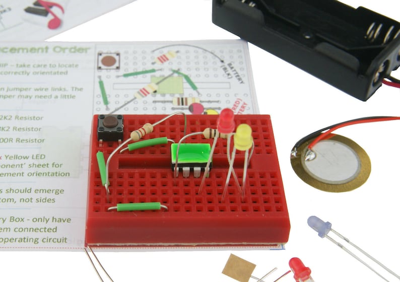 May include: A red breadboard with electronic components, including a green LED, a yellow LED, a red LED, a button, resistors, and jumper wires. The breadboard is connected to a battery pack and a piezo buzzer.