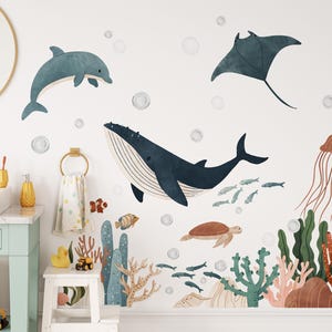May include: Wall decals featuring an underwater scene with a blue whale, dolphin, manta ray, and jellyfish. The design includes coral, fish, and bubbles, creating a whimsical ocean theme. The decals are designed for a child's room or bathroom.