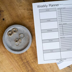 Könnte beinhalten: Ein Wochenplaner in grau-weißem Design. Der Planer hat Abschnitte für jeden Tag der Woche sowie Abschnitte für wichtige Aufgaben, die nächste Woche, Notizen und das Wochenende. Der Planer liegt auf einer Holzoberfläche.