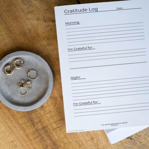 Peut inclure: Un journal de gratitude imprimable blanc avec le texte "Gratitude Log" en haut et des lignes pour écrire. Le journal est divisé en deux sections : "Matin" et "Soir". Chaque section a un espace pour écrire "Je suis reconnaissant pour : ". Le journal est sur une surface en bois avec un petit plat gris contenant des boucles d'oreilles dorées.