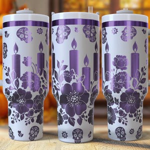 May include: Three white travel mugs with purple floral, skull, and candle designs. Each mug has a white handle and lid with a purple rim. The mugs are on a wooden surface with a blurred background of pumpkins and fall foliage.