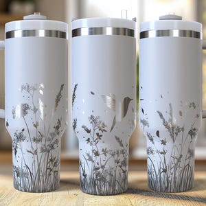 May include: Three white insulated tumblers with silver accents and handles. Each tumbler features a wraparound design of silver-colored wildflowers and a hummingbird. The tumblers have a lid and a handle for easy carrying.