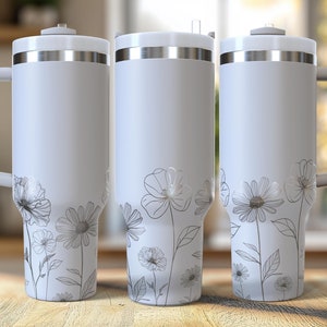 Puede incluir: Tres vasos térmicos blancos con detalles plateados y asas. Cada vaso presenta un diseño floral plateado. Los vasos están sobre una superficie de madera, con un fondo borroso de una cocina y una ventana.