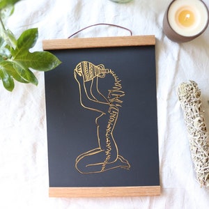 May include: A black and gold print of a woman pouring water from a pitcher. The woman is kneeling and the water is flowing from the pitcher over her head. The print is framed with a wooden frame.