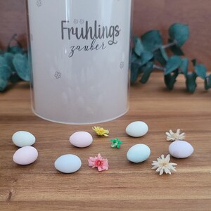 Könnte beinhalten: Ein klarer, zylindrischer Behälter mit dem Text "Frühlingszauber" und kleinen floralen Designs. Pastellfarbene Eier und Trockenblumen sind auf einer Holzoberfläche verstreut, mit Eukalyptuszweigen im Hintergrund.
