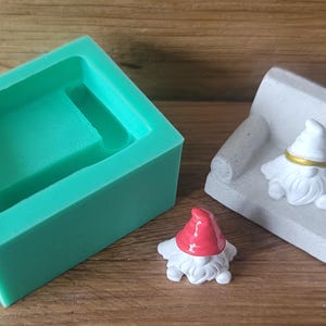 Könnte beinhalten: Eine türkisfarbene Silikonform mit quadratischer Form, neben einer kleinen weißen Zwergfigur mit roter Mütze und einer weißen Zwergfigur, die auf einem weißen Sofa sitzt. Die Zwergfiguren haben weiße Bärte.