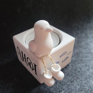 Könnte beinhalten: Ein weißer Kerzenhalter mit einer Vogelfigur mit geschlossenen Augen und weißen Stiefeln. Der Halter trägt den Schriftzug "AHOI" und blaue Fischillustrationen. Dekorativer Gegenstand.