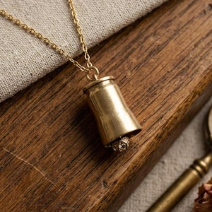 May include: A gold-toned metal pendant shaped like a spent bullet casing, suspended from a matching gold-toned chain. The pendant features a small, textured metallic sphere attached to the bottom opening of the casing, creating a bell-like appearance.