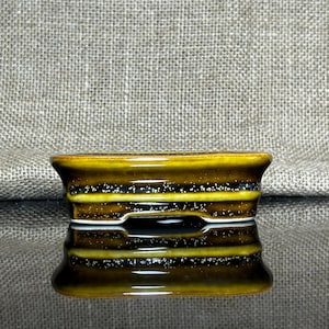 Puede incluir: Una pequeña maceta rectangular de cerámica para bonsái con acabado brillante. La maceta es principalmente marrón con detalles en amarillo y negro, y un patrón moteado blanco. La maceta se refleja en la superficie inferior.