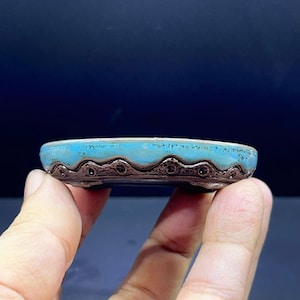 Può includere: Un piccolo vaso per bonsai in ceramica poco profondo con un bordo smaltato blu turchese vivace che sfuma in una base marrone scuro testurizzata. Il lato esterno presenta un motivo decorativo a onde smerlate con piccole rientranze circolari.
