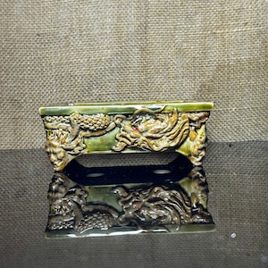 May include: A rectangular, green ceramic container with ornate dragon and face relief carvings. The container is reflected in the dark surface below. The carvings are detailed with gold and red accents. The background is a textured, tan fabric.