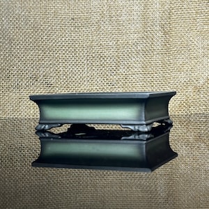 Puede incluir: Una maceta rectangular para bonsái con un borde negro mate y un panel central de color verde oscuro. El recipiente cuenta con cuatro pequeños pies ornamentados en las esquinas y un borde liso ligeramente ensanchado. El diseño es simple, funcional y simétrico.