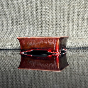 Puede incluir: Una maceta rectangular de bonsái de color rojo y marrón con un acabado brillante y un borde ligeramente elevado. La maceta tiene cuatro patas y se refleja en una superficie negra.