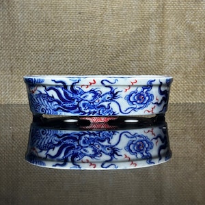 Könnte beinhalten: Eine rechteckige, weiße Keramikschale mit einem blauen Drachen- und Blumendesign. Der Drache ist mit komplizierten Details dargestellt, und die Blumen sind stilisiert. Rote Akzente werden im gesamten Design verwendet. Die Schale spiegelt sich auf einer dunklen Oberfläche.