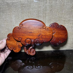 May include: A wooden cloud-shaped sculpture with a rich, reddish-brown hue. The sculpture features three connected cloud-like forms with intricate carved details, including swirling patterns. The wood grain is visible, adding to its natural aesthetic.