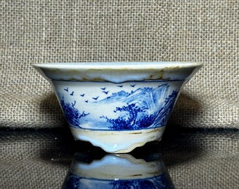 May include: A small, white ceramic bowl with a flared rim. The bowl features a blue and white landscape design with mountains, trees, and flying birds. The bowl's exterior has a slightly irregular, textured edge.