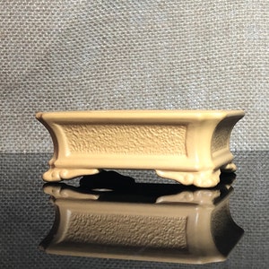 May include: A rectangular, light brown ceramic bonsai pot with a textured surface and four feet. The pot is sitting on a black reflective surface.