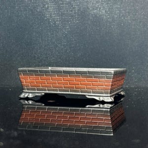 Puede incluir: Una maceta rectangular con un patrón de ladrillos en filas alternas de gris oscuro y marrón rojizo. El recipiente tiene una parte superior abierta y ancha, y se apoya sobre cuatro pies pequeños, ornamentados y curvos en la base.