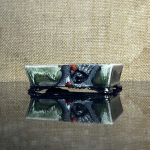 May include: A rectangular ceramic bonsai pot featuring a dark green and white drip glaze finish. The center of the front side displays a sculpted black relief detail with small red and white accents, supported by four small, dark, ornate feet.
