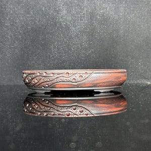 Może przedstawiać: Niska, owalna doniczka ceramiczna do bonsai z wykończeniem w kolorze ciemnego brązu i czerwonawego brązu. Zewnętrzna strona posiada teksturowany, wypukły wzór z wirujących linii i małych okrągłych wgłębień wokół boku naczynia.