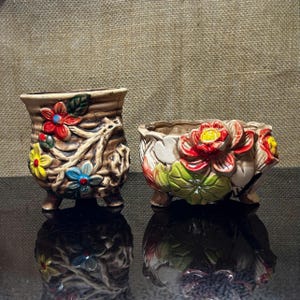 Op de afbeelding: Twee keramische bloempotten met bloemmotieven. De ene is een hoge, getextureerde pot met kleurrijke bloemen en wijnranken. De andere is een lage, brede pot met rode, gele en groene bloemen. Beide potten hebben kleine voetjes.