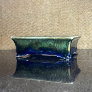 May include: A rectangular bonsai pot with a glossy, glazed finish. The ceramic pot has a gradient of dark green and blue hues, with a slightly flared rim and small feet. The pot is reflected in the surface below.