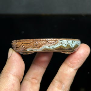 Pode incluir: Um vaso de cerâmica para bonsai, circular e raso, com um exterior castanho texturizado que apresenta padrões circulares esculpidos e uma faixa ondulada vidrada em azul claro ao longo da borda superior. O vaso assenta em pequenos pés.