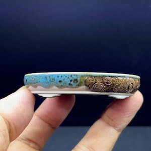Könnte beinhalten: Ein flacher, ovaler Bonsai-Topf aus Keramik mit einer leuchtend türkisblauen Glasur am oberen Rand, die in eine strukturierte, dunkelbraune Oberfläche mit kreisförmigen Prägemustern an der Seite übergeht. Der Boden hat kleine Füße.