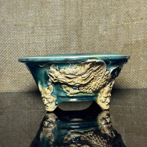 Puede incluir: Una maceta de bonsái de cerámica color verde azulado con una escultura en relieve de un dragón que envuelve el exterior. La maceta se apoya sobre tres patas, cada una esculpida con la forma de un rostro detallado. El esmalte es brillante.