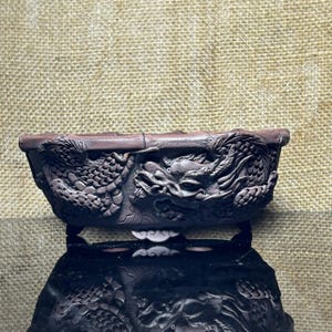 Könnte beinhalten: Ein dunkelbrauner, rechteckiger Bonsai-Topf mit detaillierten Reliefschnitzereien eines Drachen, der sich um die Außenwände windet. Das Gefäß hat einen dicken, strukturierten Rand und steht auf kleinen, verzierten Füßen mit ausgeprägten Schuppen.