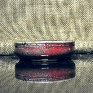 Puede incluir: Un cuenco pequeño y redondo de cerámica con interior rojo y exterior oscuro y moteado. El borde es de un color más claro, posiblemente blanco o crema. El cuenco se refleja en una superficie brillante.