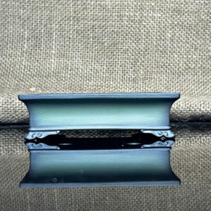 Puede incluir: Una caja decorativa rectangular de color verde azulado con un interior oscuro. La caja presenta diseños curvos y ornamentados en los laterales. La caja está hecha de un material sólido y tiene una superficie reflectante.