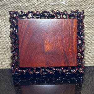 Puede incluir: Un expositor cuadrado de madera marrón rojizo con un borde tallado de color marrón oscuro. El expositor presenta una superficie lisa y pulida y detalles intrincados y calados alrededor de los bordes, mostrando la veta natural de la madera.