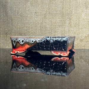 Puede incluir: Una maceta rectangular de cerámica para bonsái con un esmalte multicolor. El exterior muestra un patrón de rejilla negro texturizado, una base naranja vibrante y un esmalte blanco estilo goteo en el borde superior. Se apoya sobre cuatro pies.