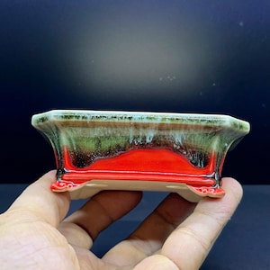 Könnte beinhalten: Ein rechteckiger Bonsai-Topf aus Keramik mit einer leuchtend roten Basis, die in eine dunkelgrüne und weiße Tropfglasur am oberen Rand übergeht. Der Topf steht auf vier kleinen, verzierten Füßen und hat Abflusslöcher am Boden.