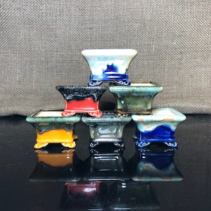May include: Six small, square bonsai pots in various colours, including blue, green, red, and yellow, with a glossy finish. The pots are stacked, highlighting their decorative design and small size. Each pot has a unique glaze.